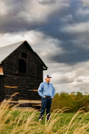 Man in blue shirt and cap standing in tall grass before a weathered black barn under stormy sky.