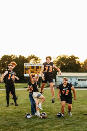 Group of five football players in black Indians jerseys celebrating on field at sunset.