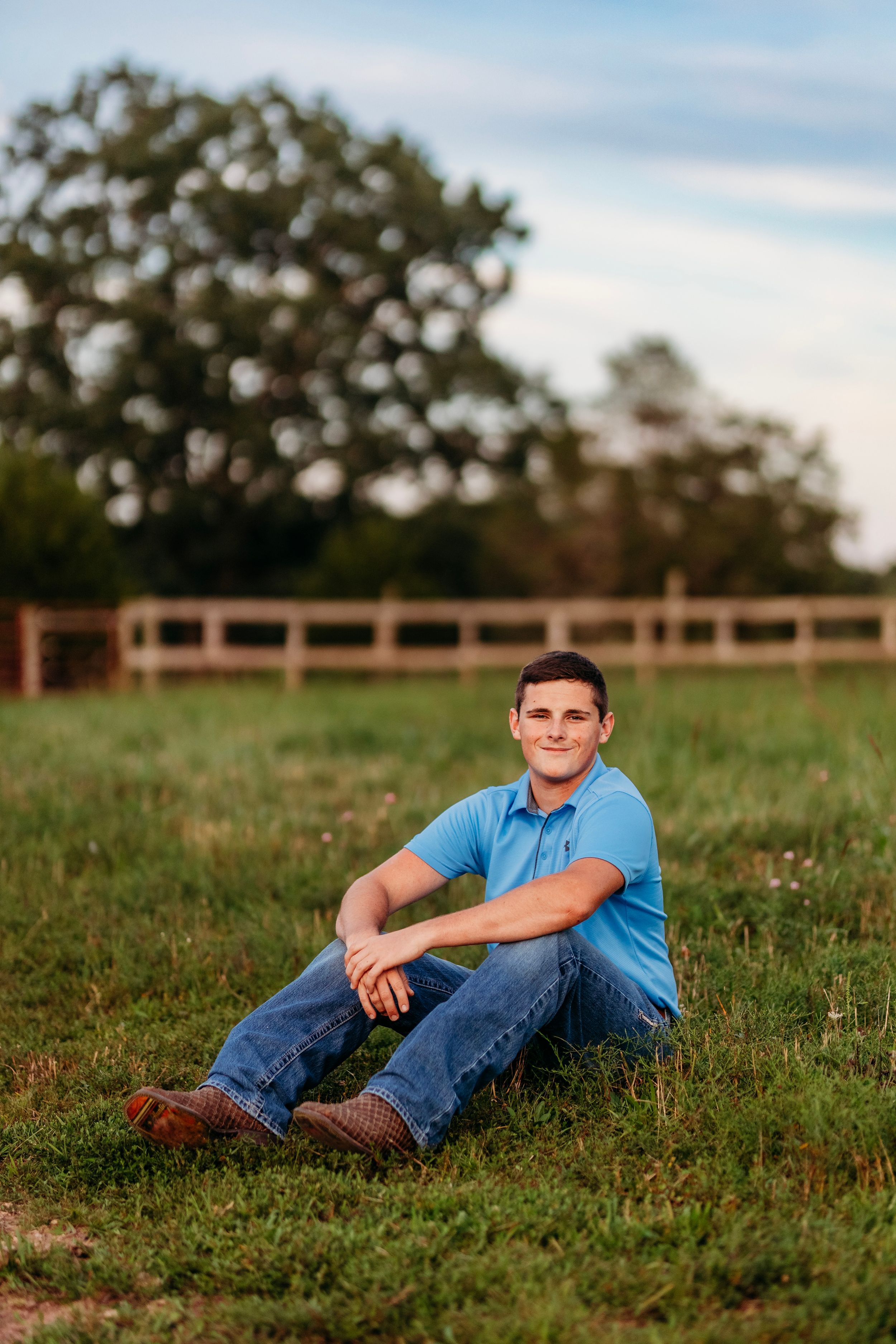 Young man in blue polo and jeans sitting relaxed on green grass with wooden fence and trees in background.