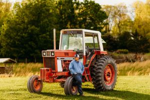Man in blue shirt leaning casually against a vintage red tractor in a sunny green field.
