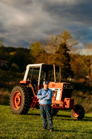 Man in blue shirt standing arms crossed before a vintage red tractor under dramatic cloudy sky.