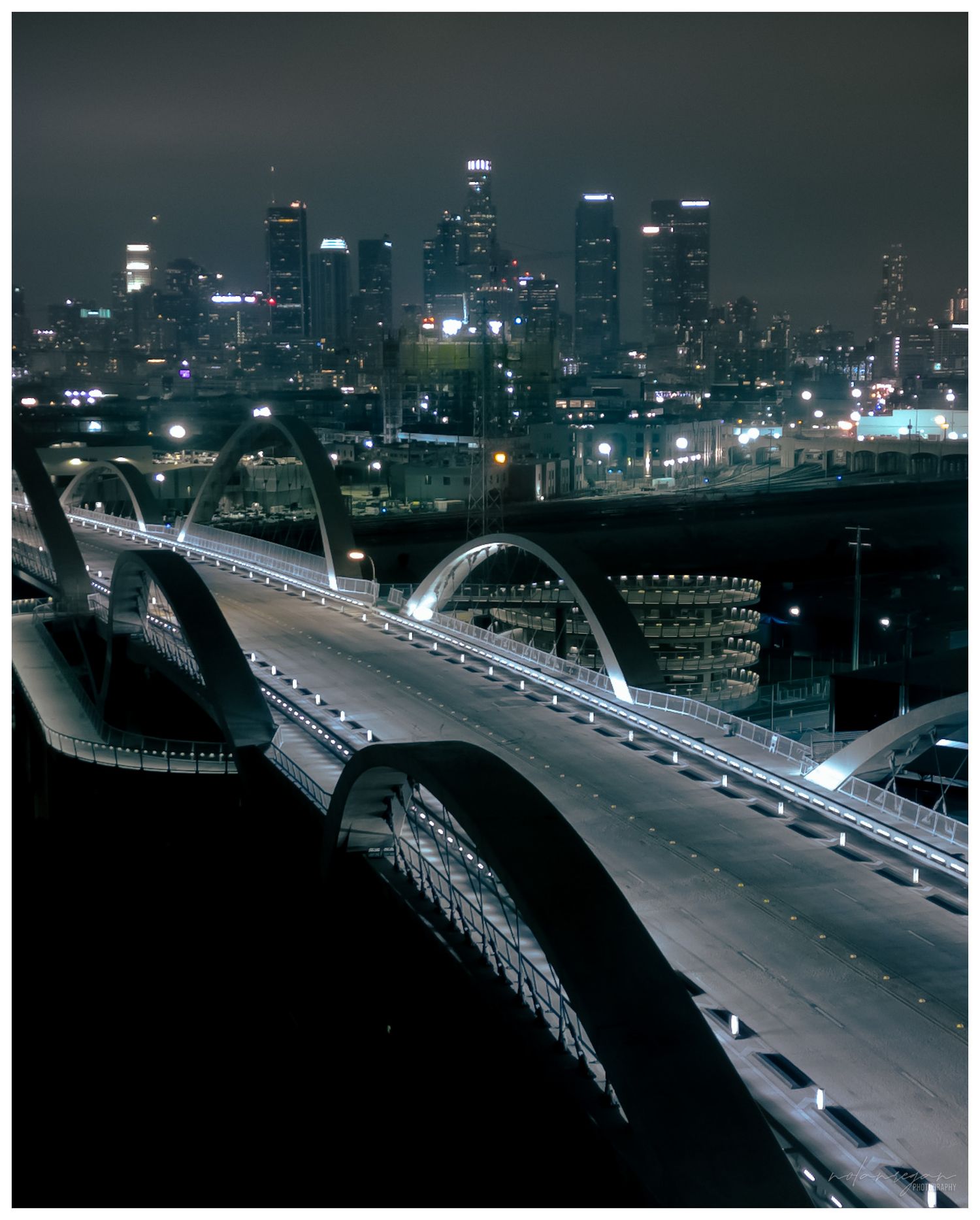 Illuminated bridge and city skyline at night with glowing lights reflecting on dark water.