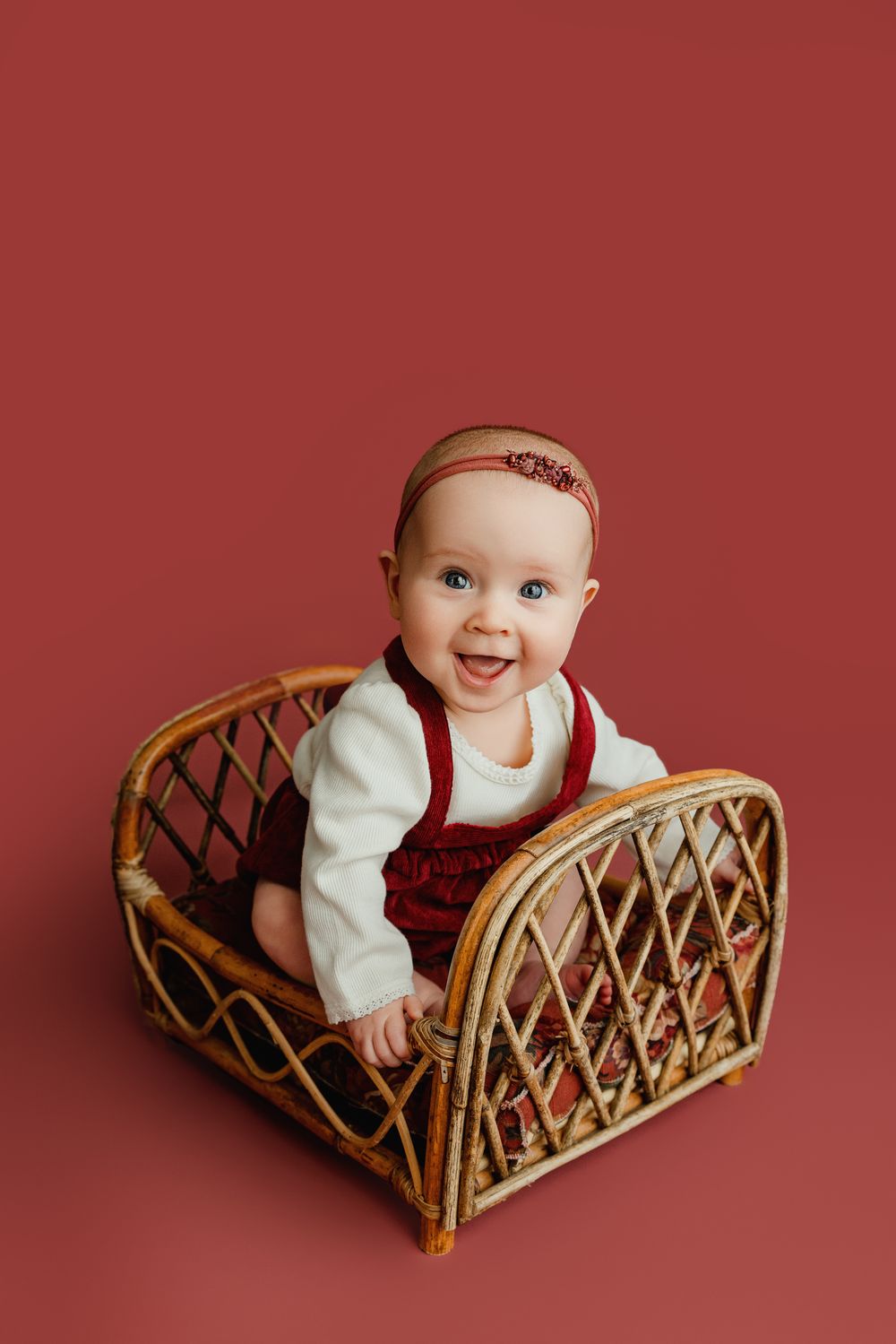 Cheerful baby sitting in rattan chair against red backdrop wearing white and burgundy outfit.