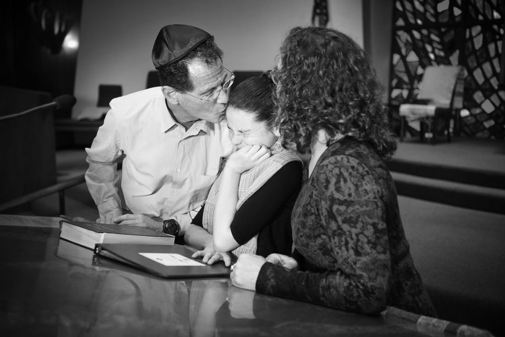 A funny and candid black and white photograph of parents kissing their daughter during her Bat Mitzvah service.