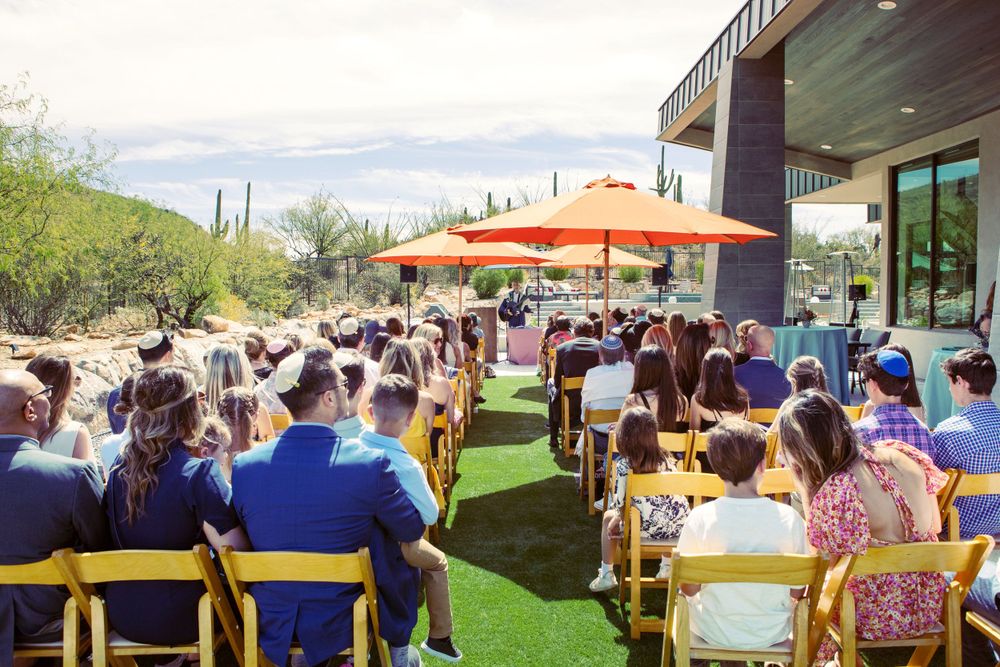A fancy home Bat Mitzvah taking place in the backyard.  Wooden chairs are placed in rows and orange umbrellas are used for shade.