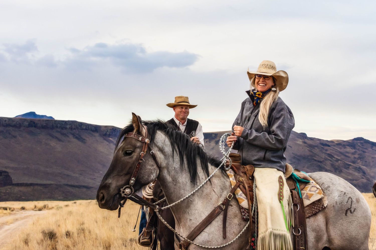 Two riders on horseback survey a rugged mountain landscape at sunset.