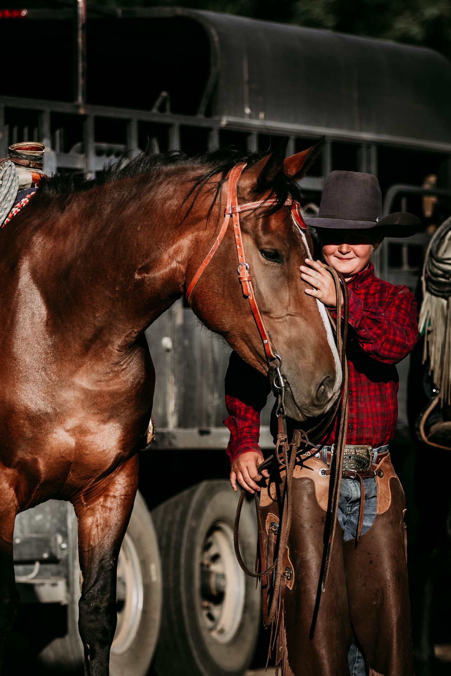 A glossy bay horse with western tack and a young cowboy stand next to a horse trailer.