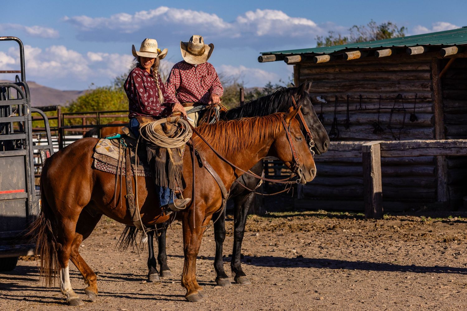 Two riders on horseback pause near wooden ranch buildings under blue skies.