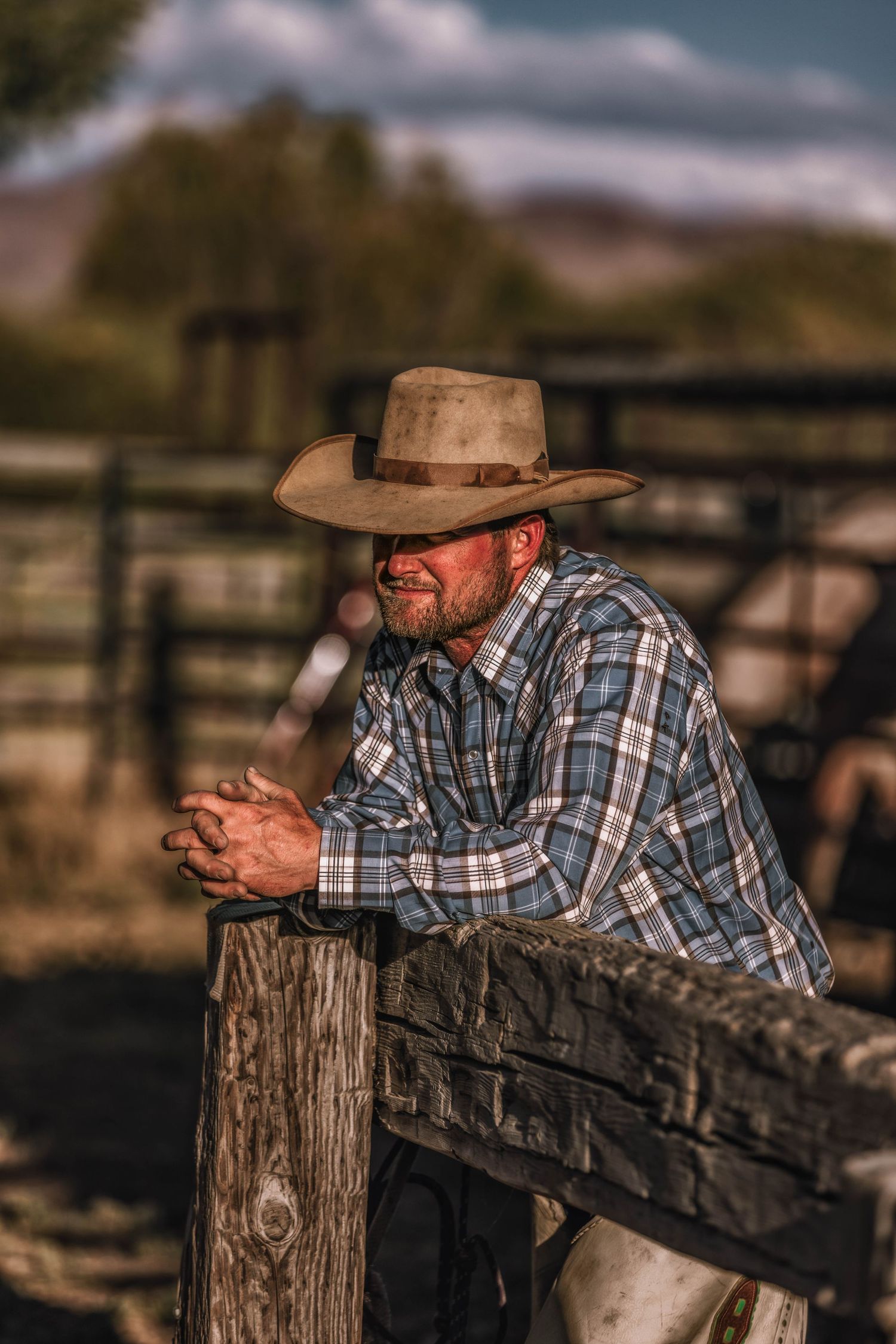 Cowboy in western wear leans on a wooden fence post at a ranch during sunset.