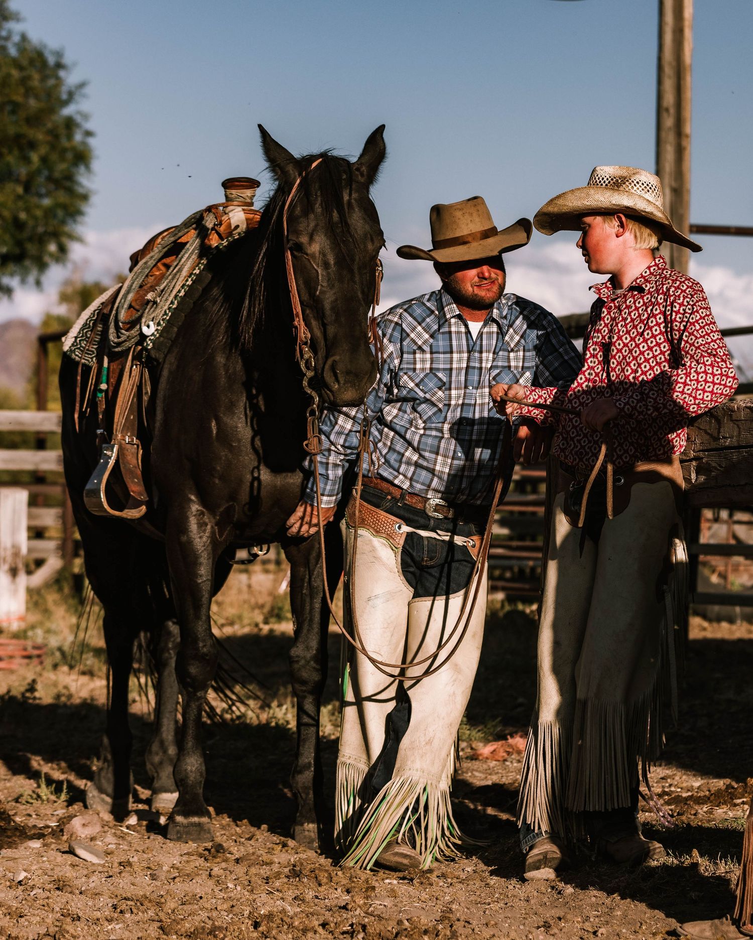 Father and son cowboys in traditional western clothing stand beside a saddled black horse.