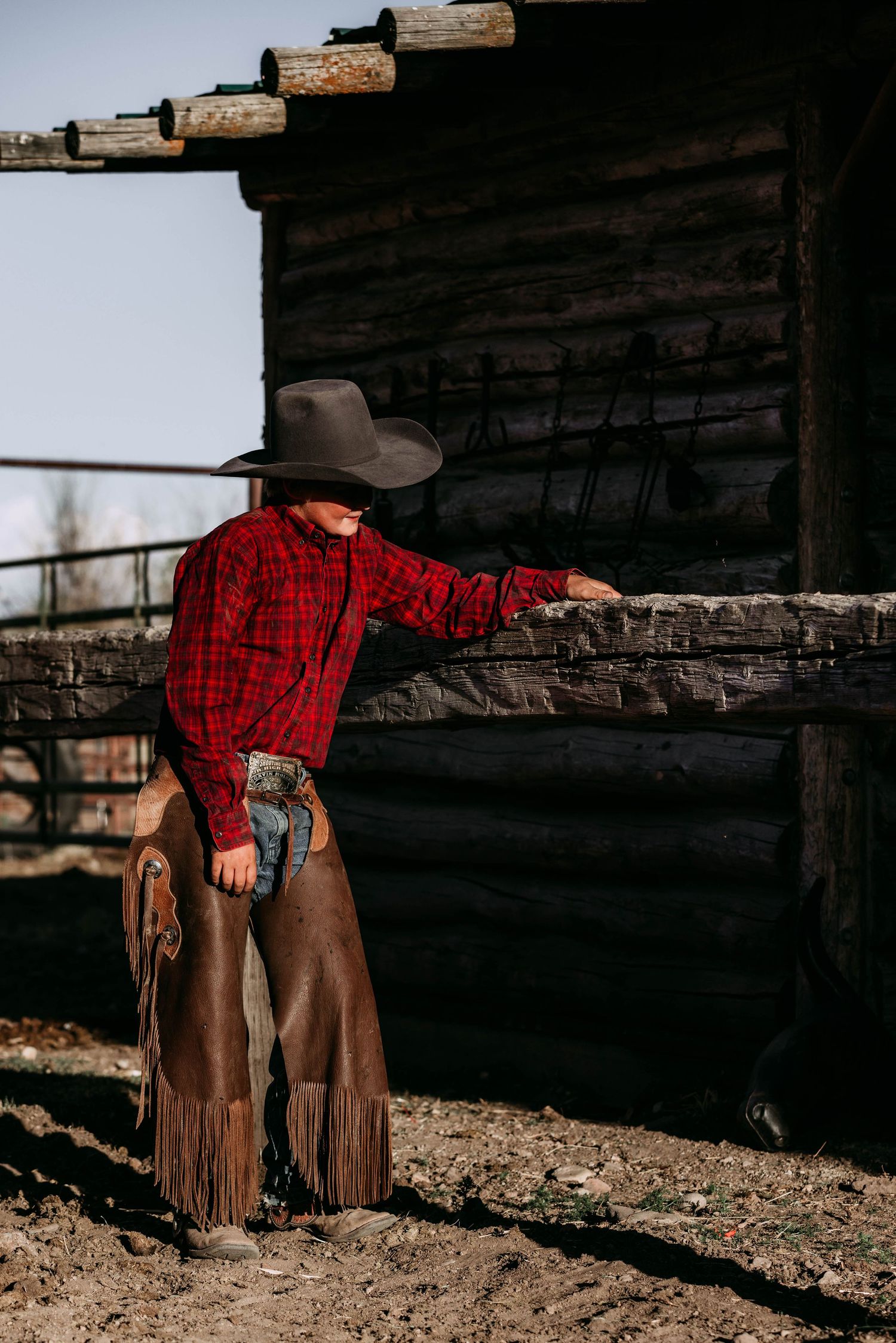 A cowboy in red plaid shirt and leather chaps leans against a rustic log cabin wall.