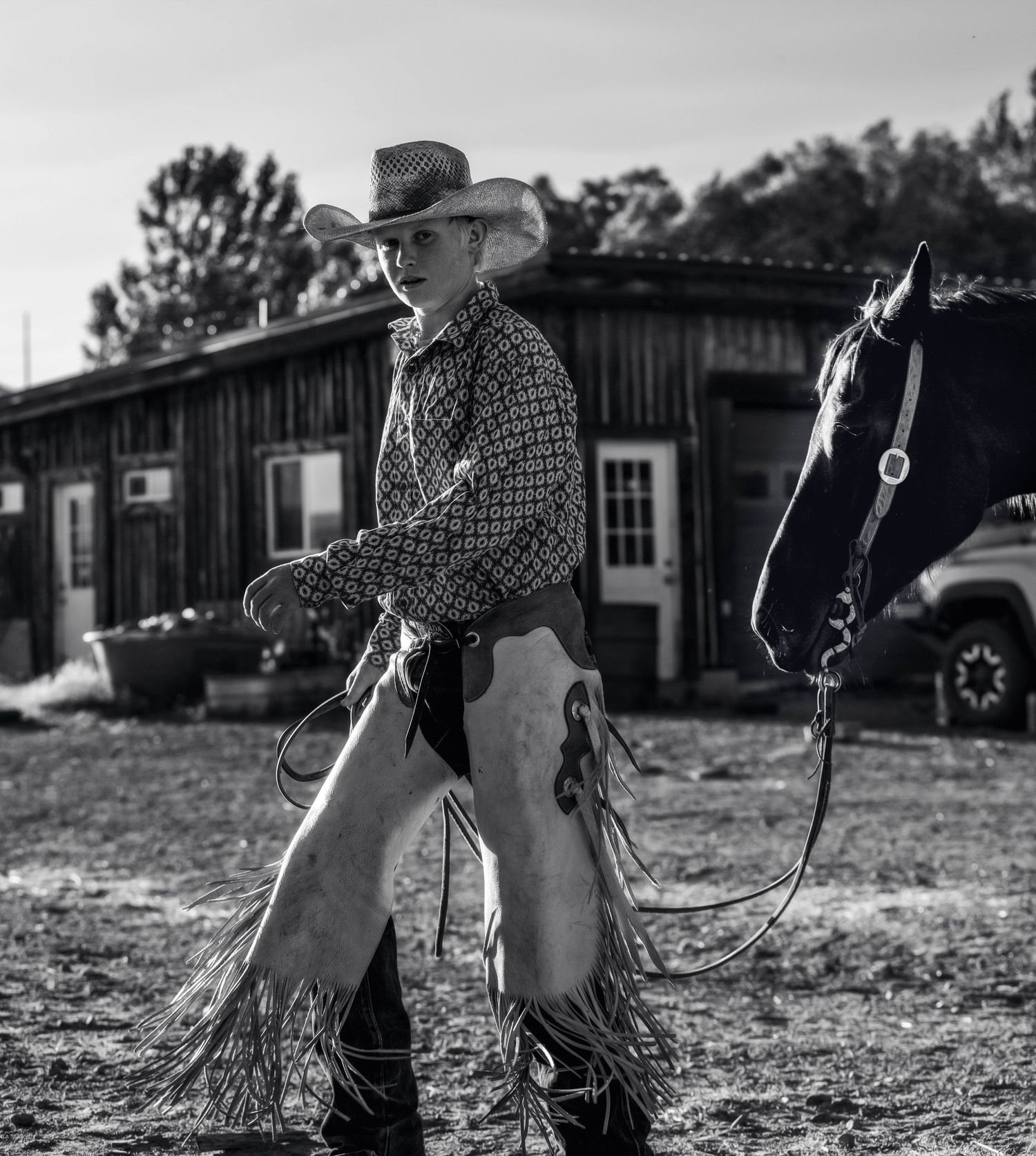 A black and white image of a cowboy in western gear leading a horse near a wooden barn.