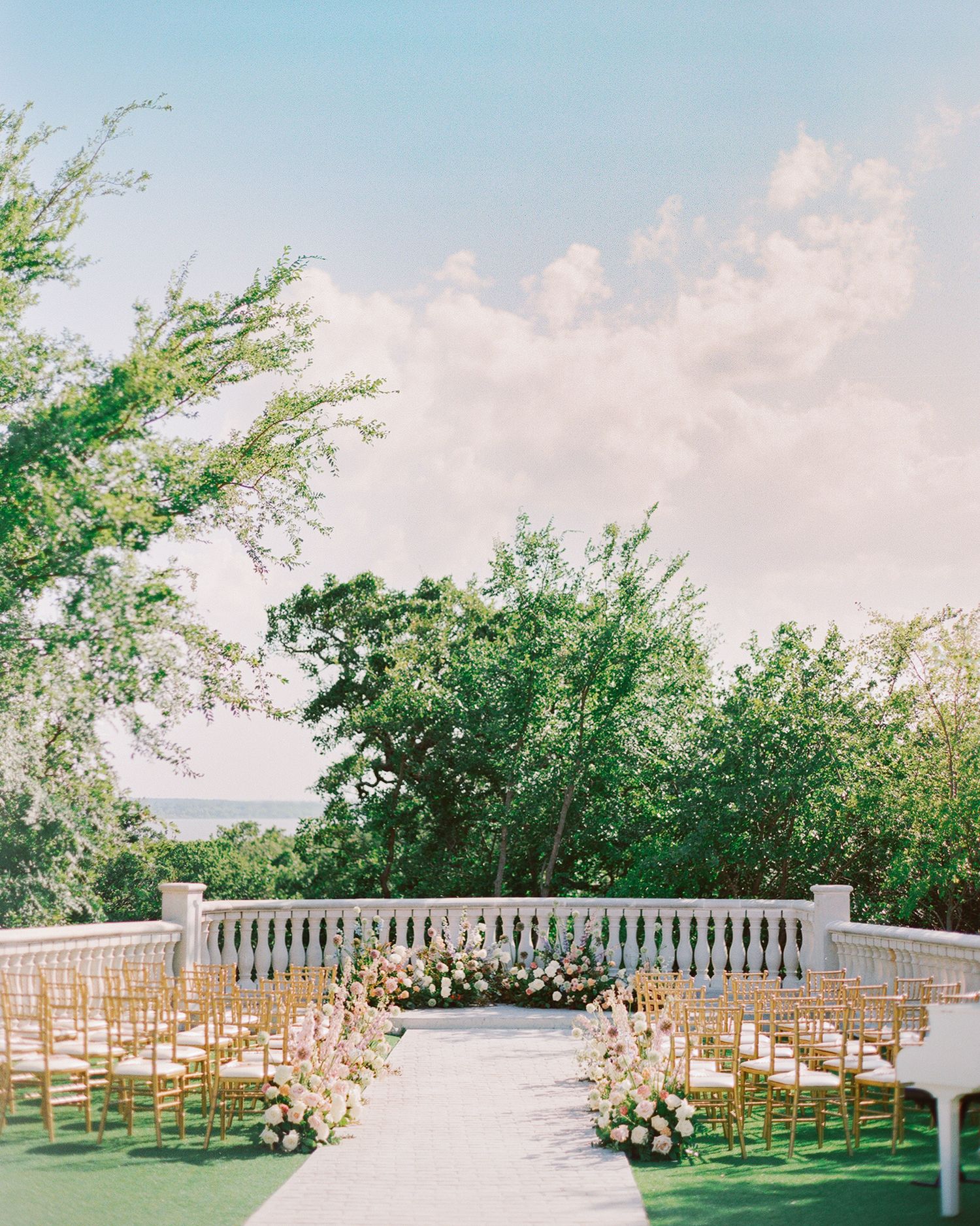Elegant outdoor wedding ceremony setup with gold chairs, floral aisle decor, white balustrade, and lush green trees under a blue sky.