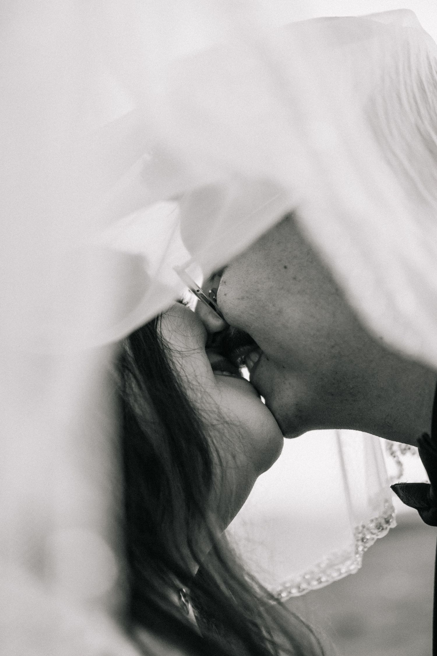 Black and white photo of a couple sharing a romantic kiss under a flowing bridal veil on their wedding day.