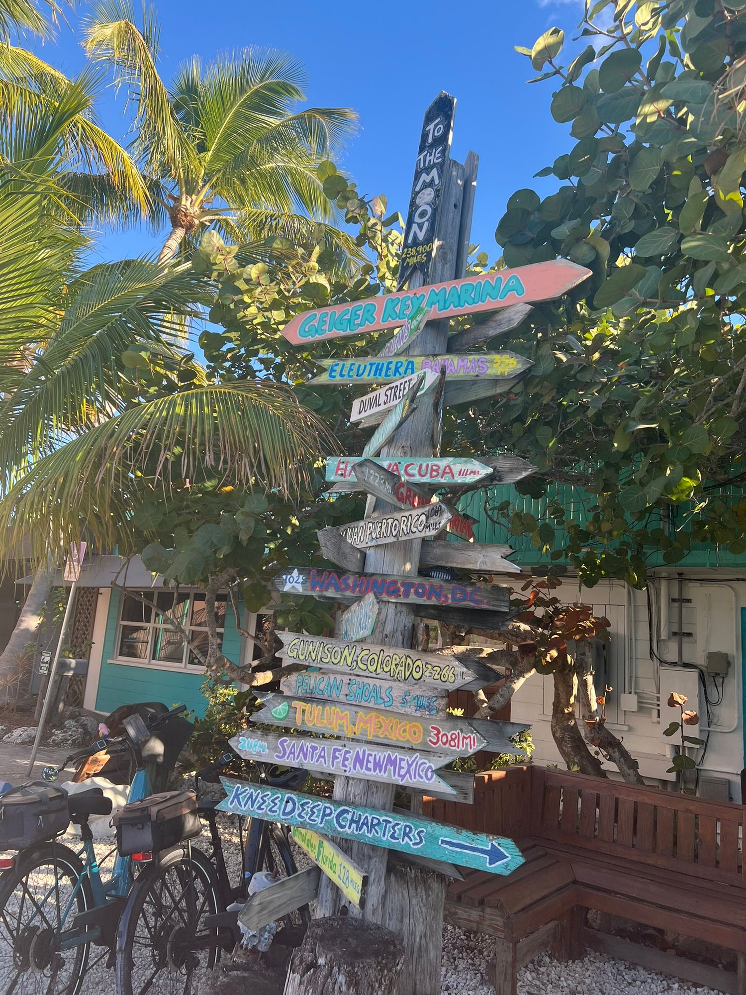 A colorful tropical directional sign post with destinations like Cuba and Colombia near palm trees and a bicycle.
