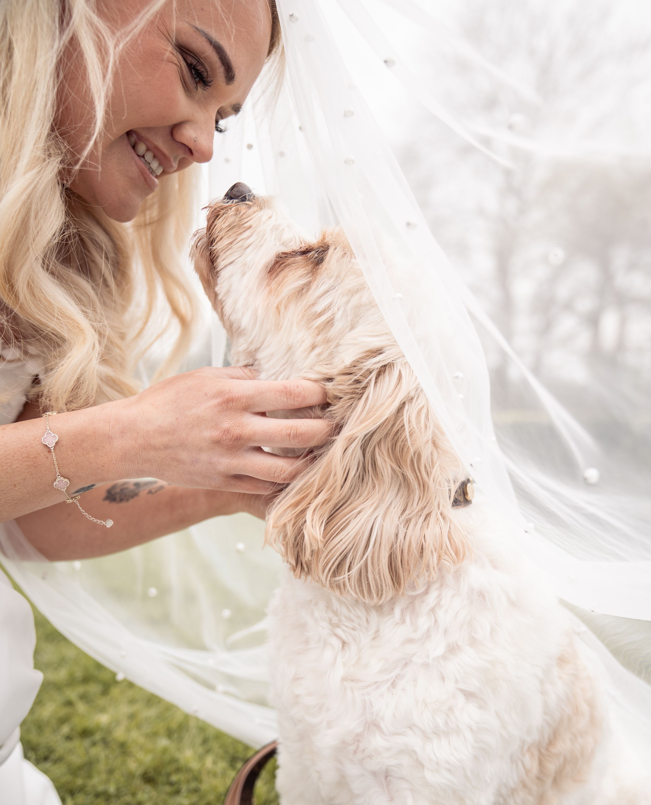 A smiling bride in a white veil tenderly holds a fluffy cream-colored dog on her wedding day outdoors.