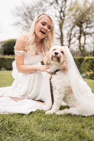 Smiling blonde bride in white dress sits on grass with a fluffy Cavapoo dog wearing her veil outdoors.