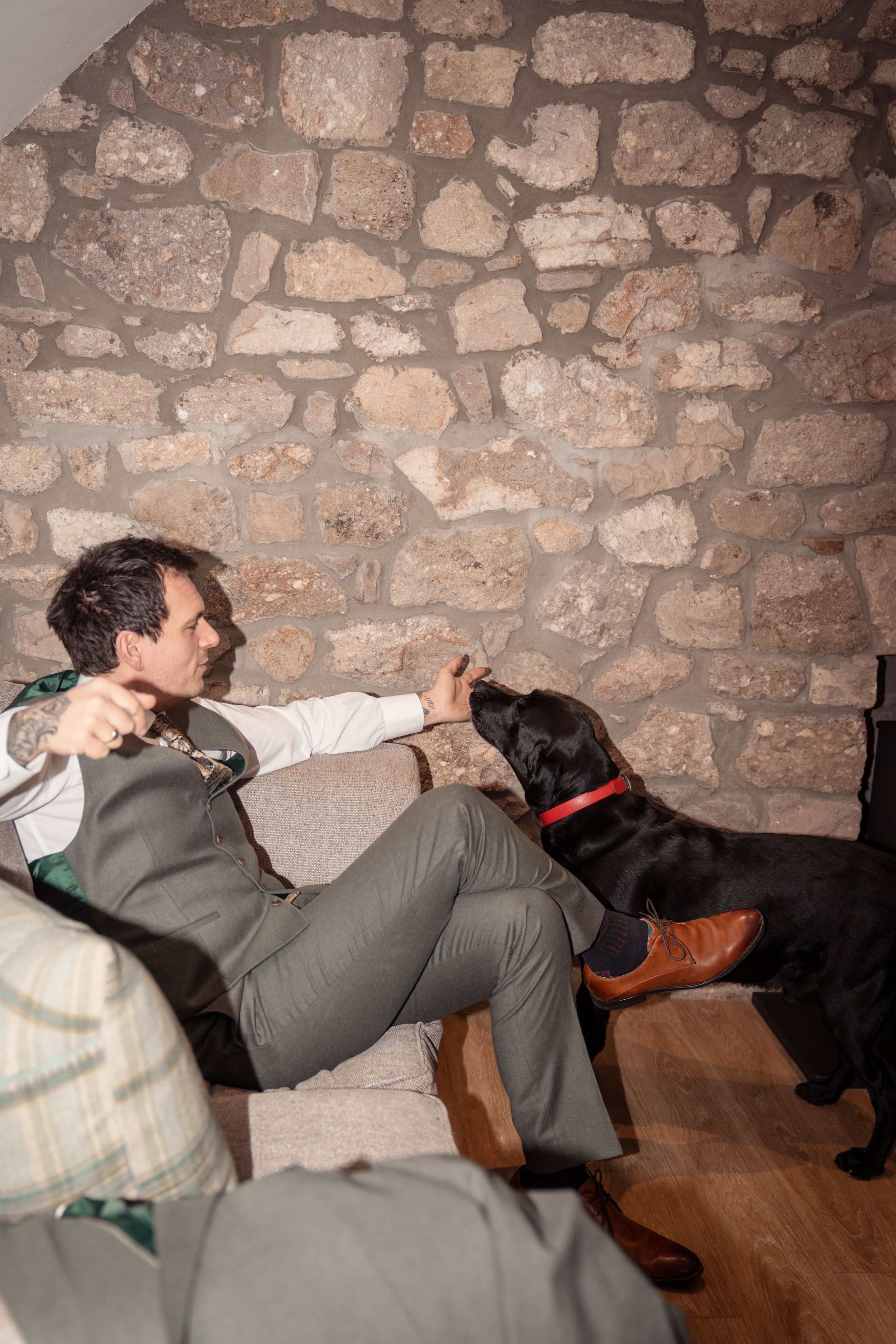 Man in suit sitting on sofa interacting with a black Labrador dog against a rustic stone wall background.