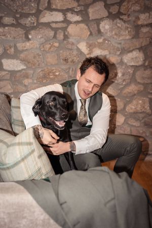 A well-dressed man in a vest and tie affectionately hugs a happy black Labrador dog on a sofa near a stone wall.