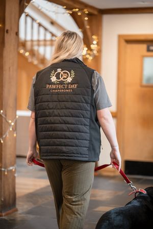 Woman in Pawfect Day Chaperones vest walking a black dog through an elegant wooden venue with fairy lights.