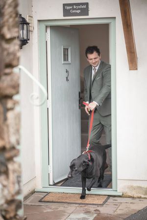 A smartly dressed man in a grey suit walks a black dog on a red leash through a cottage doorway.