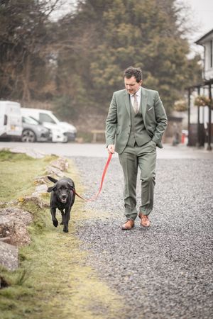 Man in green suit walking black Labrador on red leash along gravel path outdoors.