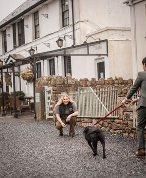 Woman crouching to greet a black dog on a red leash outside a white country inn on a gravel courtyard.