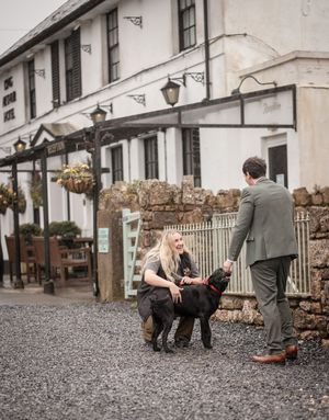 A woman crouches to pet a black Labrador dog outside a traditional white pub building with hanging baskets.