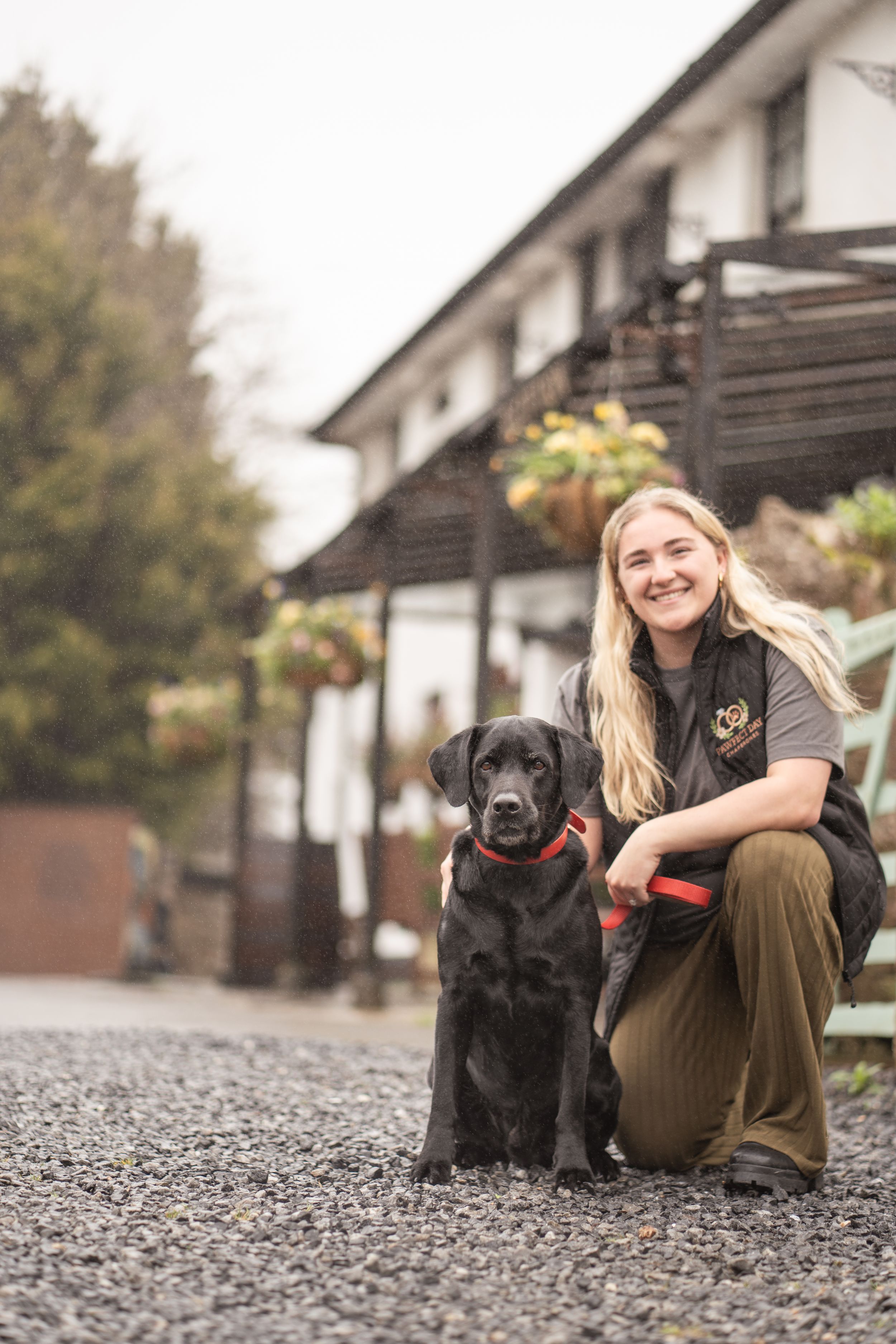 A smiling blonde woman crouches beside a black Labrador on a gravel path outside a white building.
