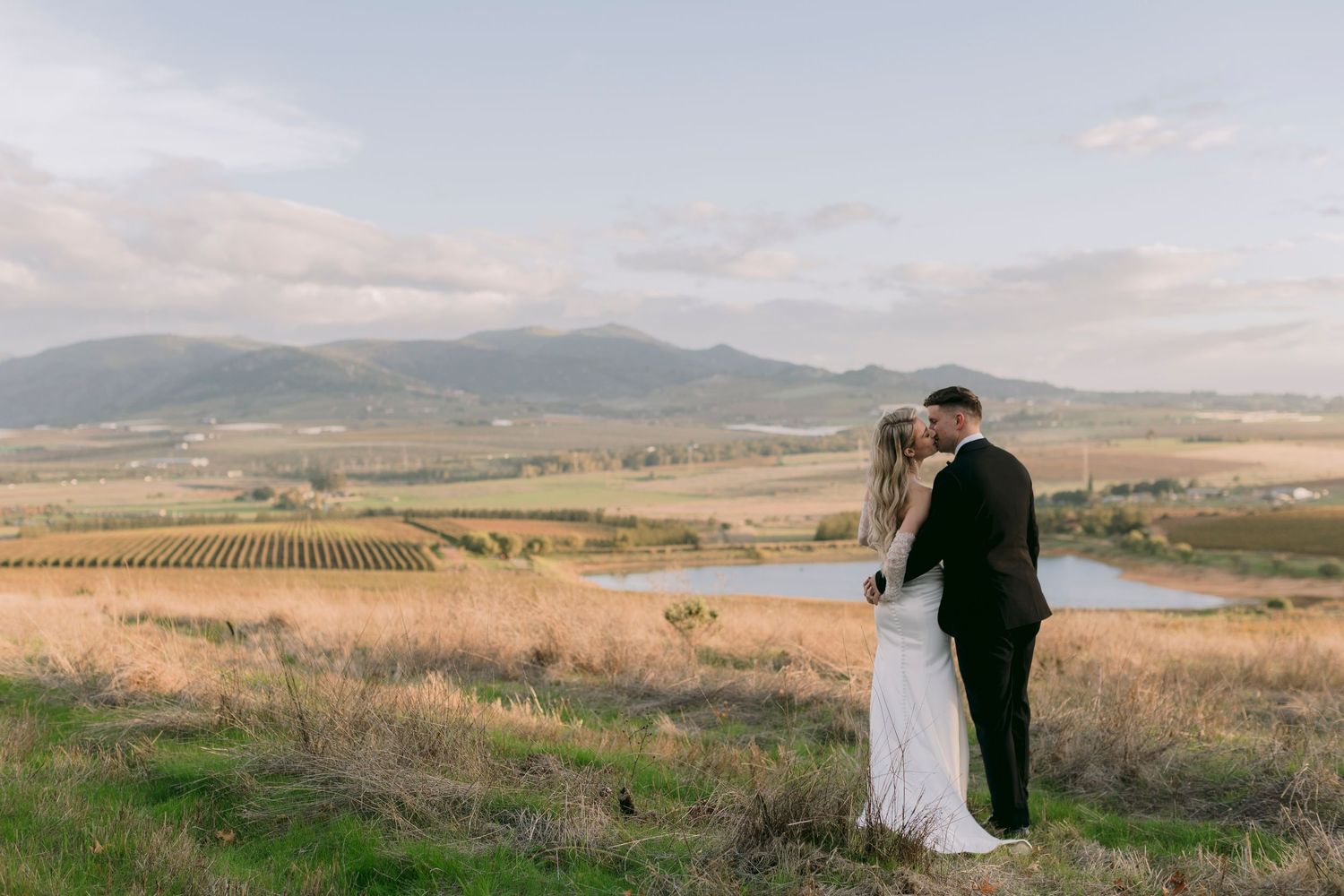 Elegant bride in off-shoulder lace wedding gown wrapped in flowing veil in soft natural light.