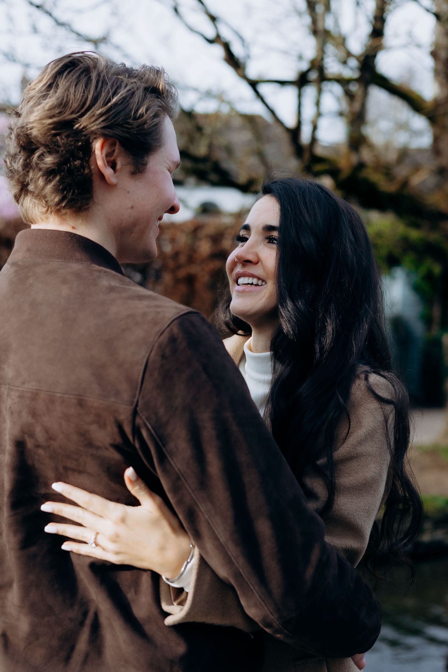Smiling dark-haired woman embraces a man in a brown suede jacket outdoors, wearing an engagement ring at their cotswolds proposal photoshoot