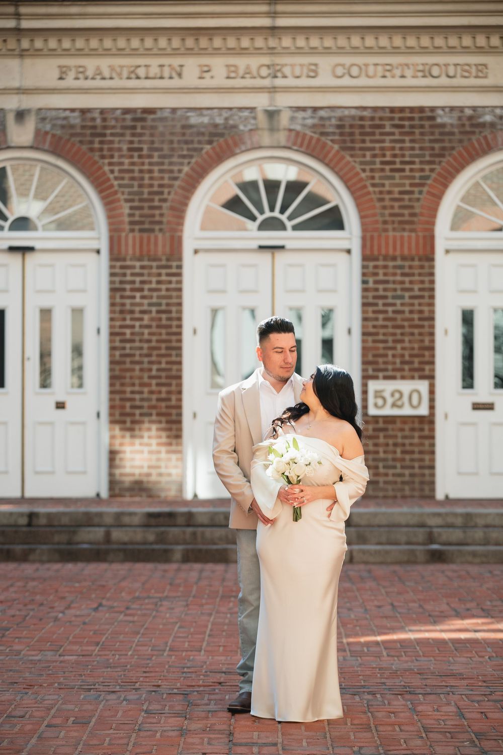 Couple in elegant attire embracing in front of Franklin P. Backus Courthouse, bride holding white bouquet.