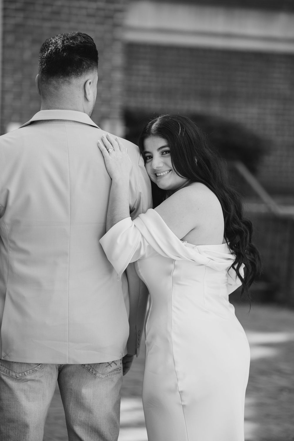 Black and white photo of a smiling woman in white dress embracing a man in a suit jacket from behind.