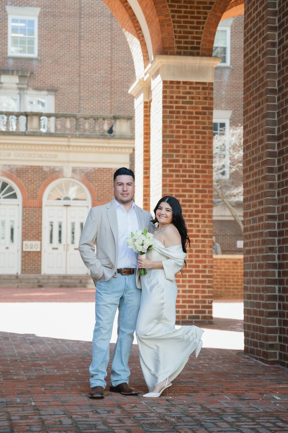 Bride in off-shoulder gown holds bouquet beside groom in tan suit under brick archway in outdoor wedding portrait.