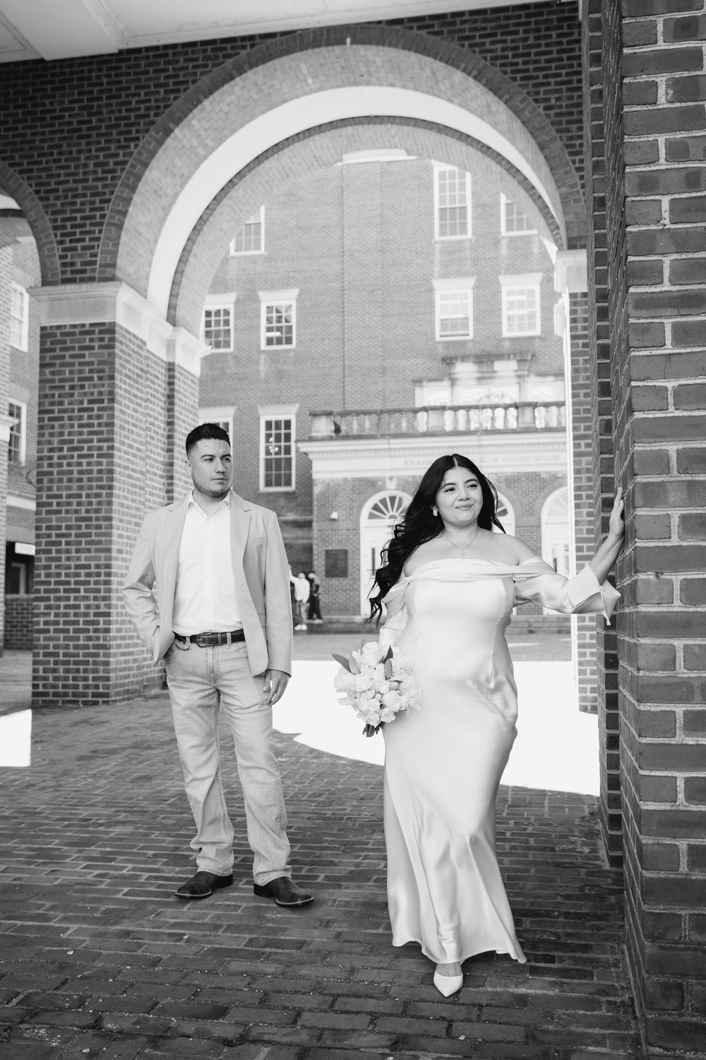 Black and white photo of bride in satin gown and groom in suit posing under brick archway.
