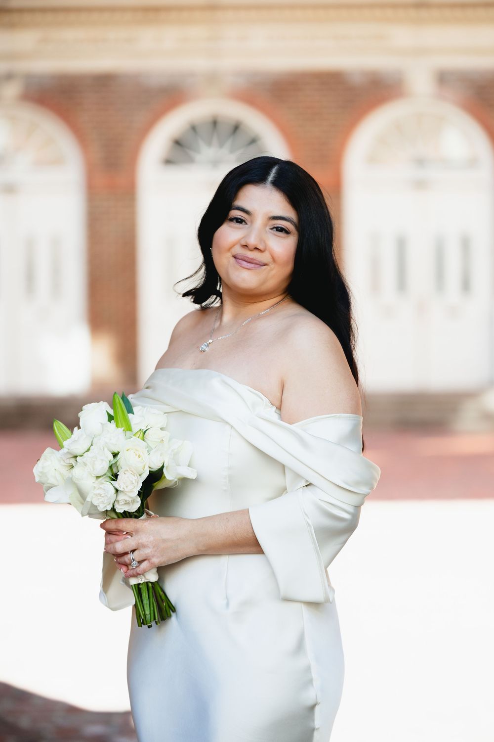 Bride in off-shoulder white gown holding white rose bouquet in front of brick building.