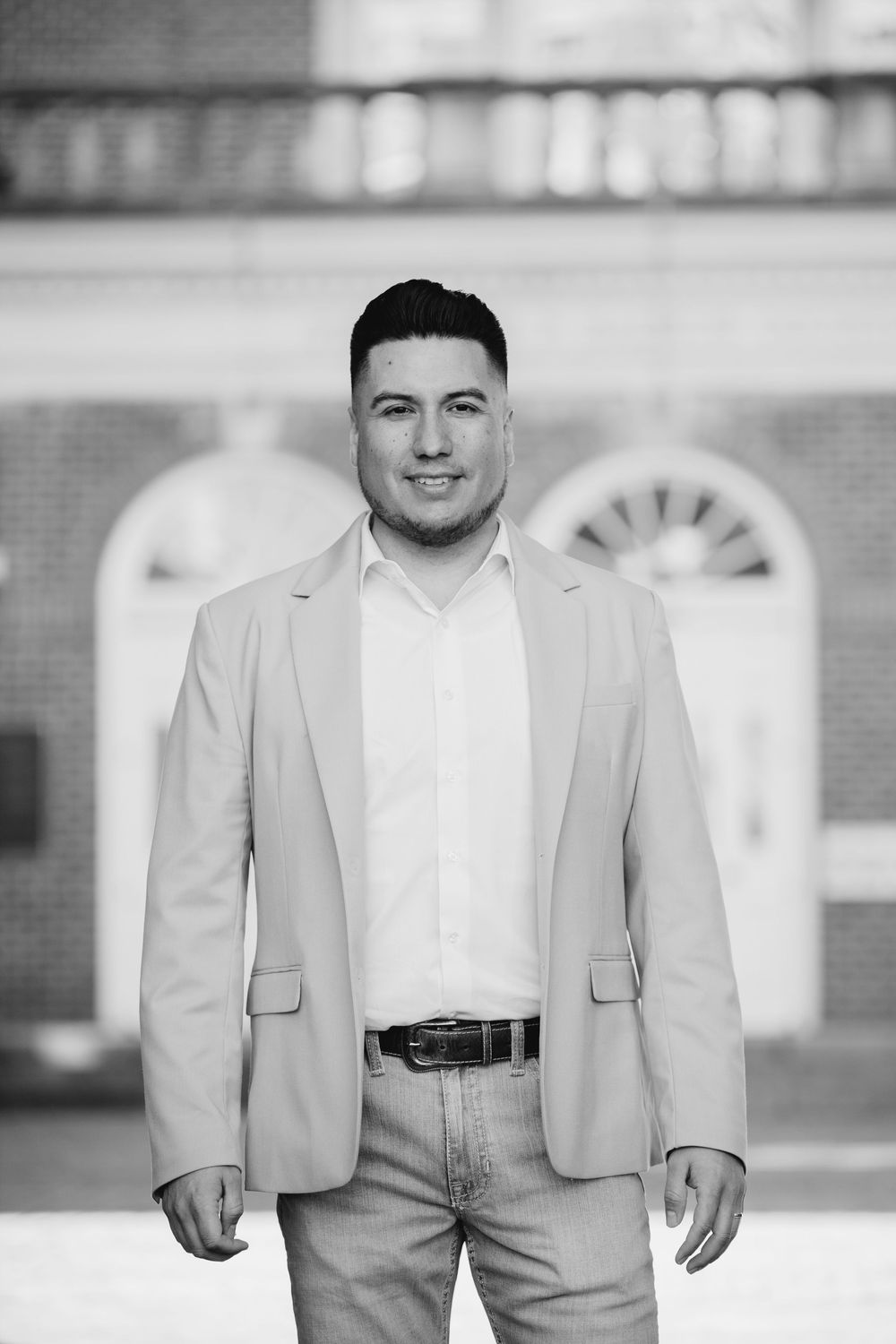 Black and white portrait of a young man in a blazer and dress shirt smiling outdoors near a brick building.
