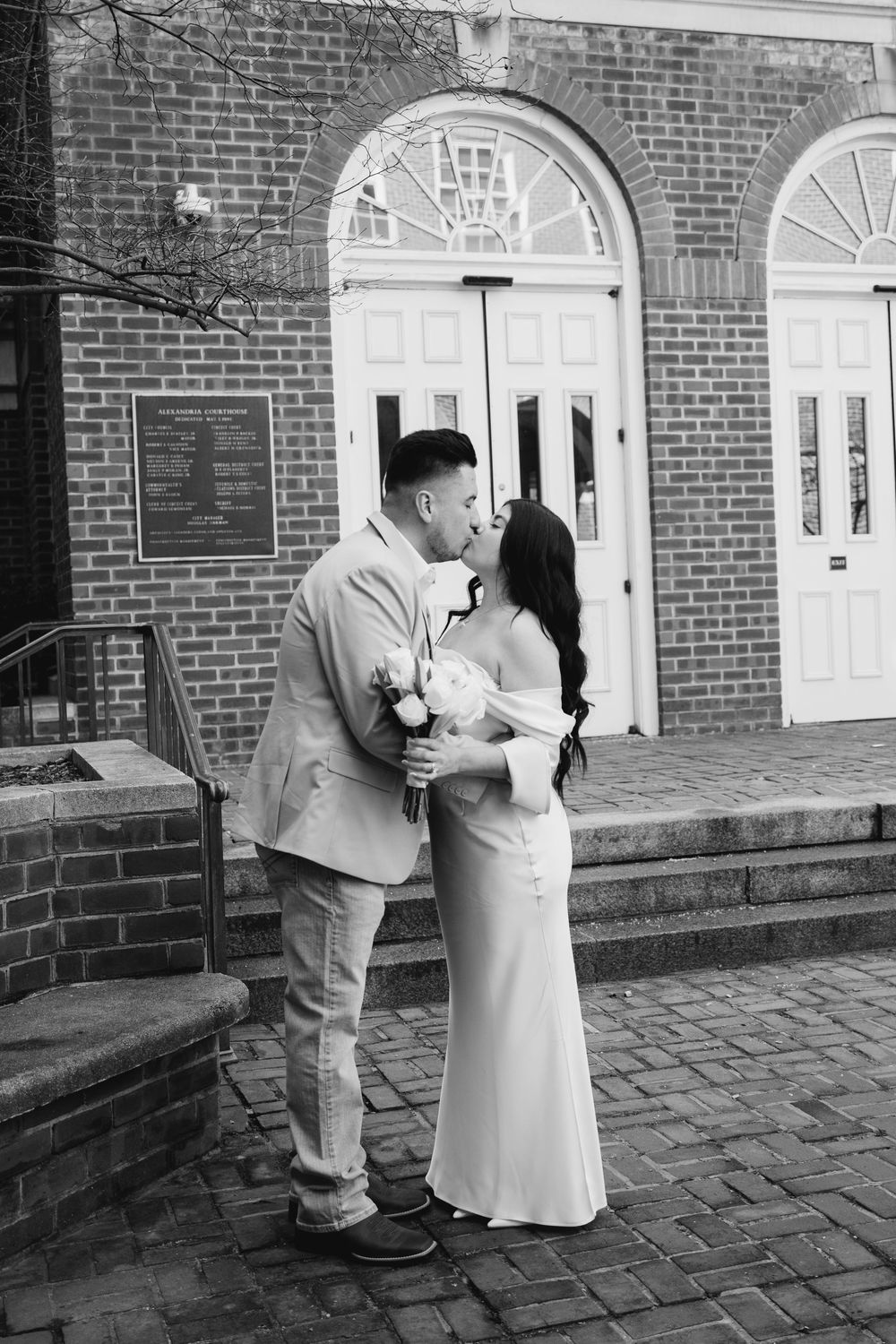 Black and white photo of a couple kissing outside a brick church, bride in white gown holding bouquet.