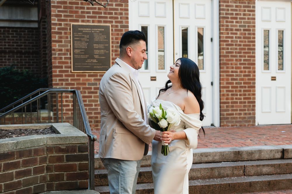 Couple in wedding attire embracing outside a brick building, bride holding white bouquet in elegant off-shoulder gown.
