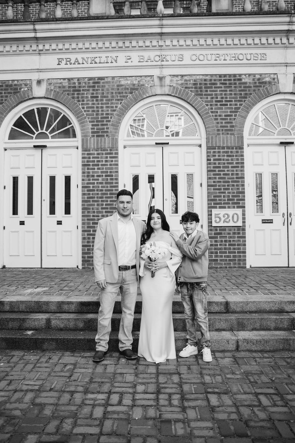Bride and groom pose with child on courthouse steps at Franklin P. Backus Courthouse in black and white wedding photo.