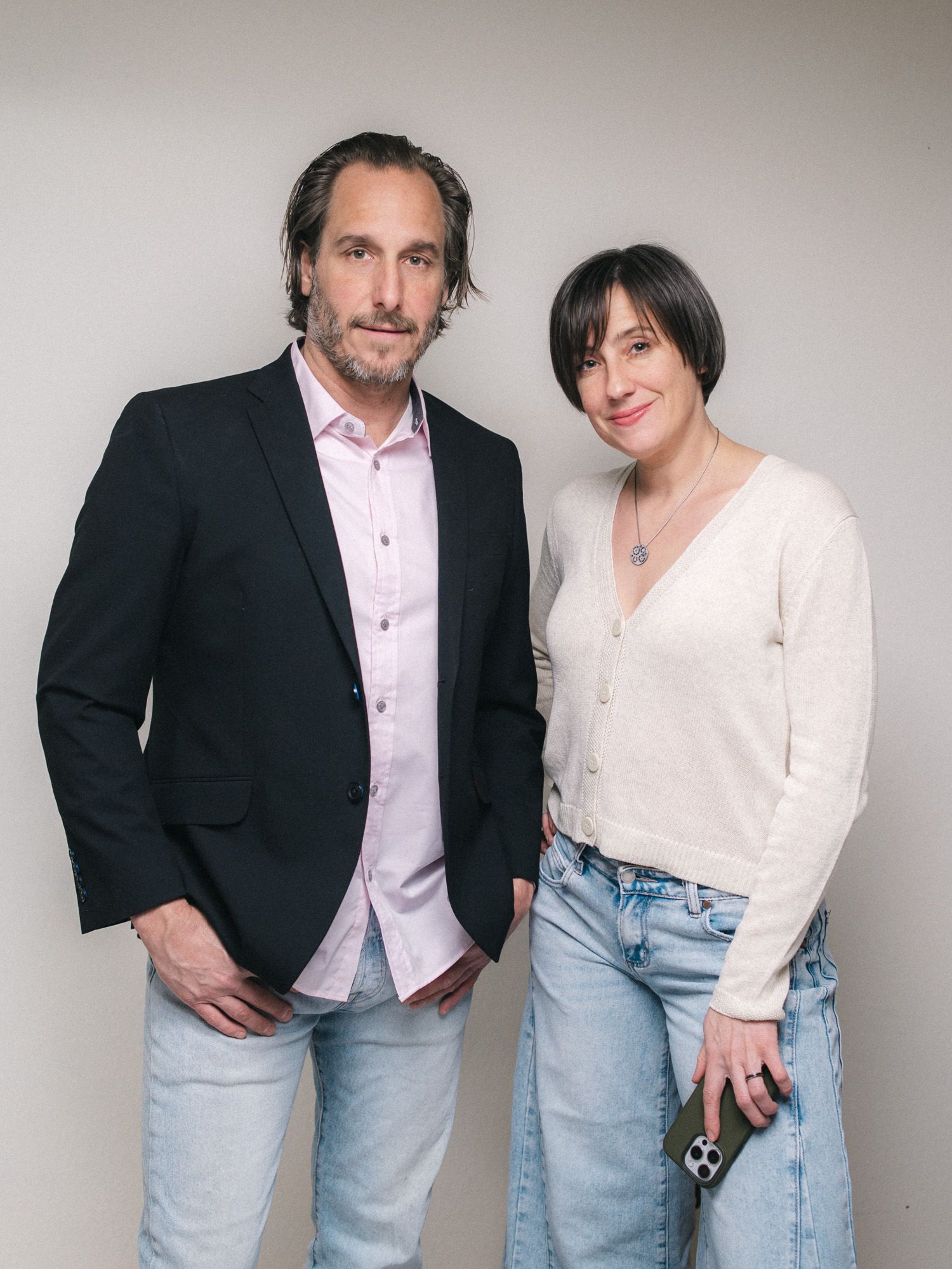 A man in a black blazer and a woman in a white cardigan pose together against a neutral background.