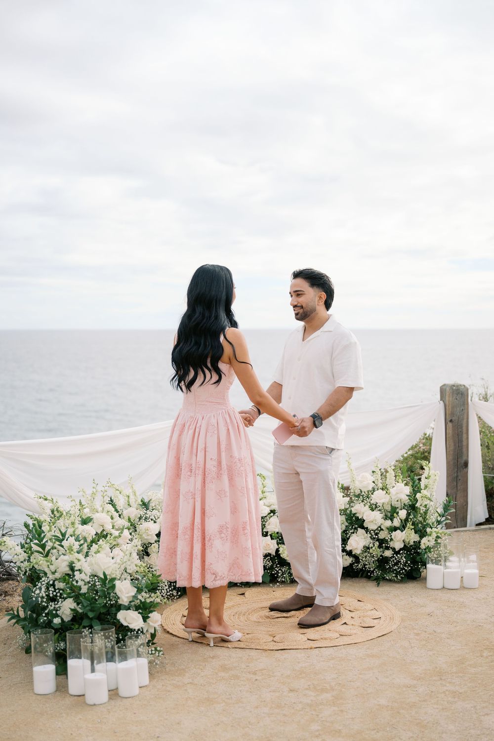 Couple holds hands during romantic cliffside proposal with white floral arrangements, candles, and ocean backdrop.