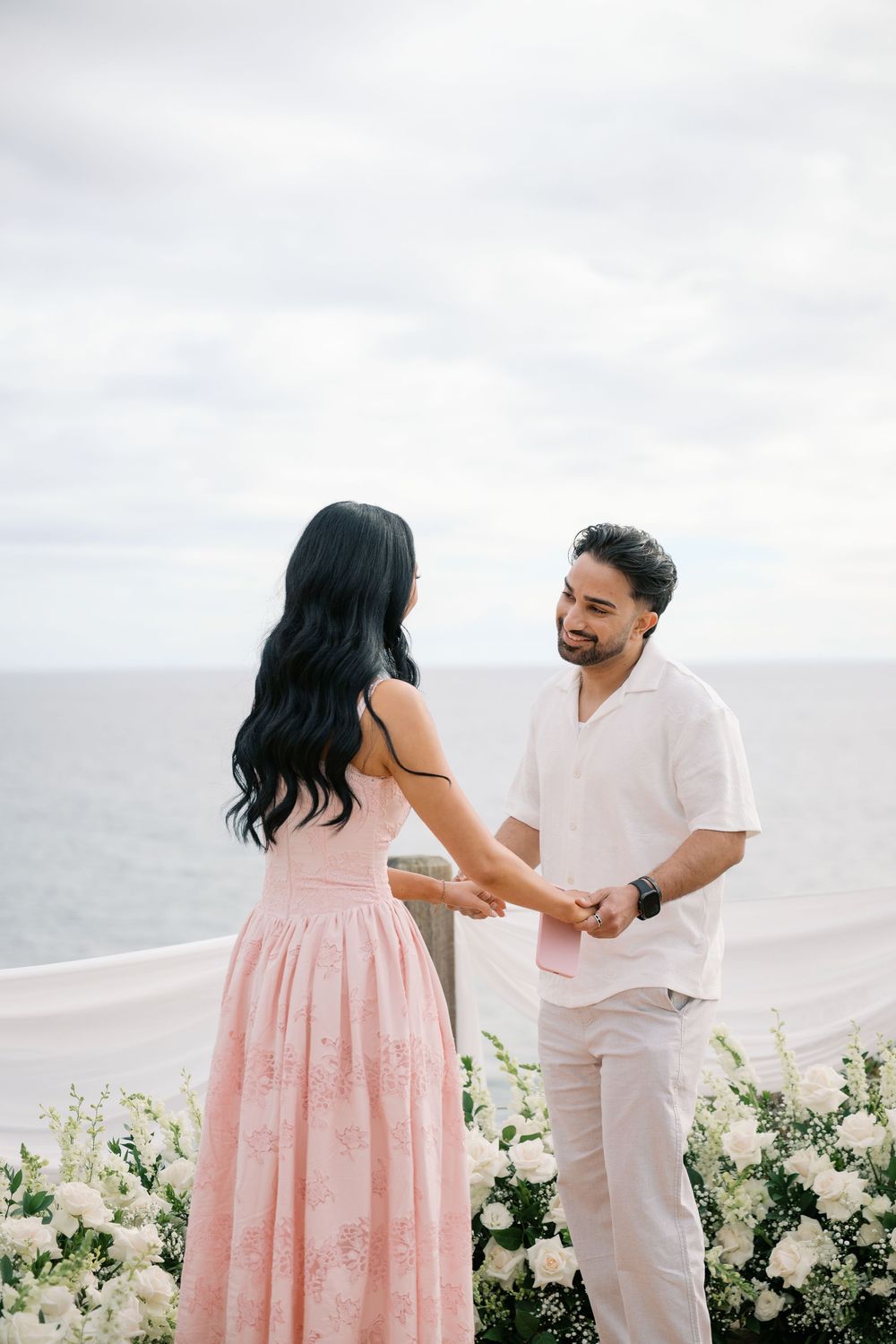 Couple holding hands at an oceanside proposal setup with white floral arrangements, woman in pink dress, man in white shirt.