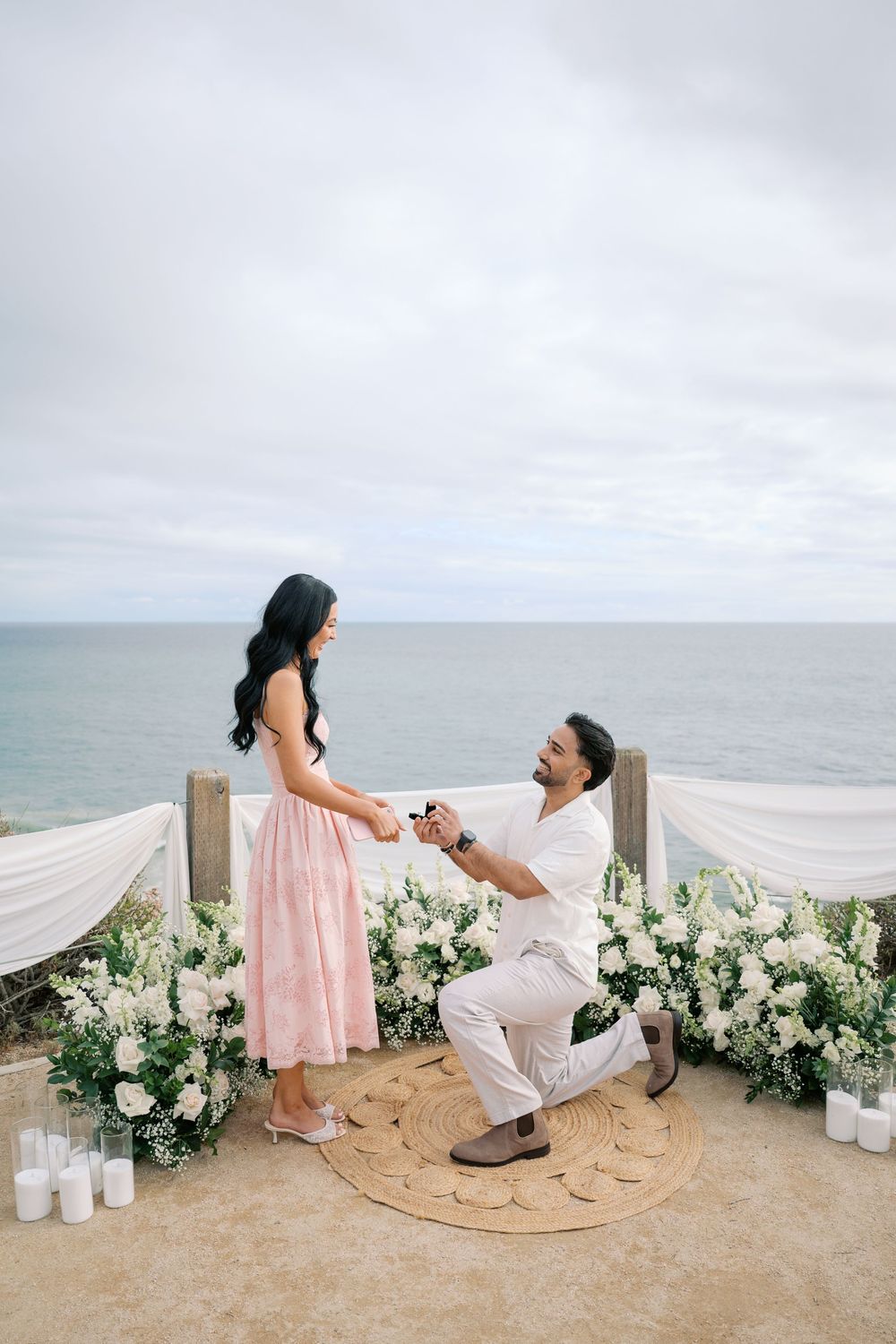 Man proposes on one knee to woman in pink dress at oceanfront floral setup with white flowers and candles.
