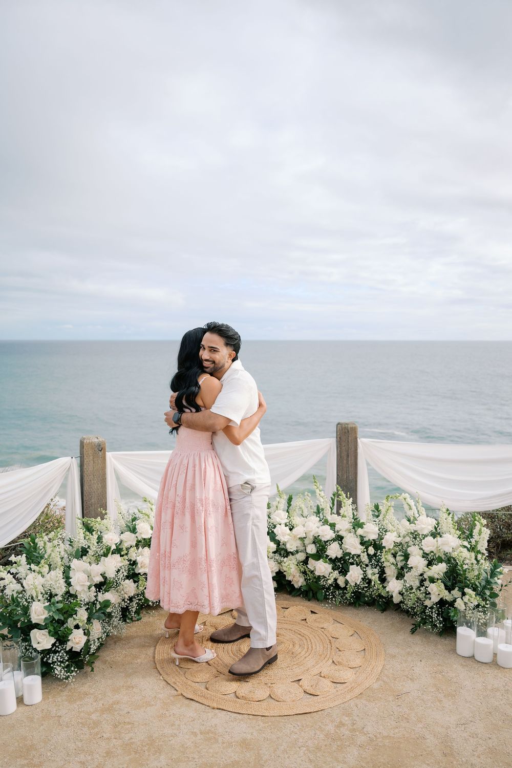 Couple embracing during romantic oceanfront proposal with white floral arrangements and candles.