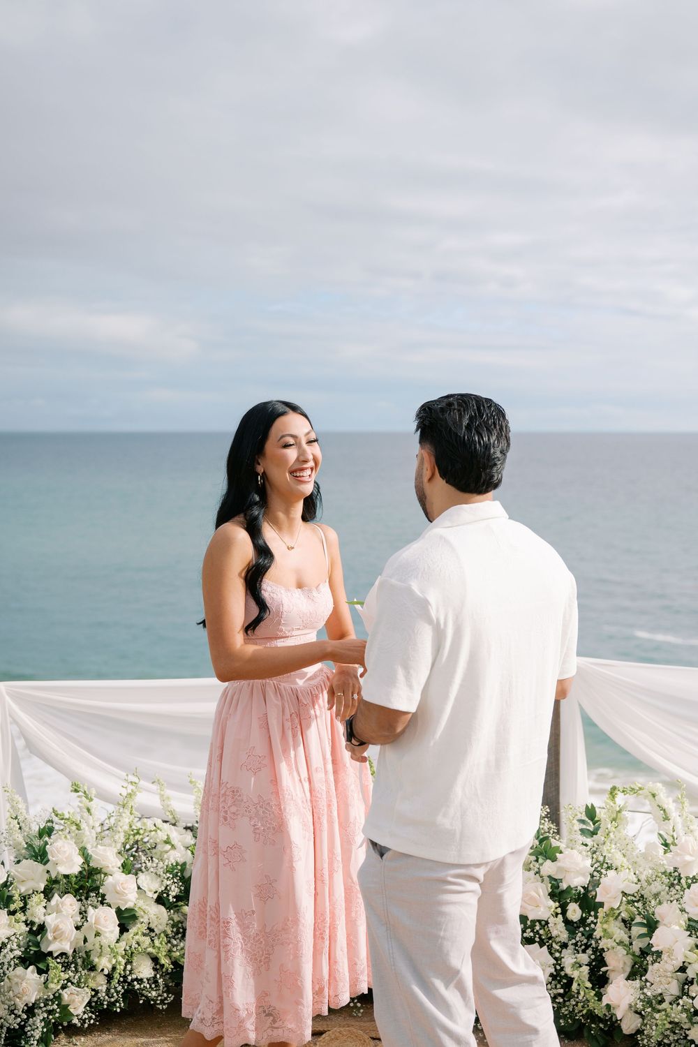 Smiling woman in pink dress holds hands with man during seaside proposal with white flowers.