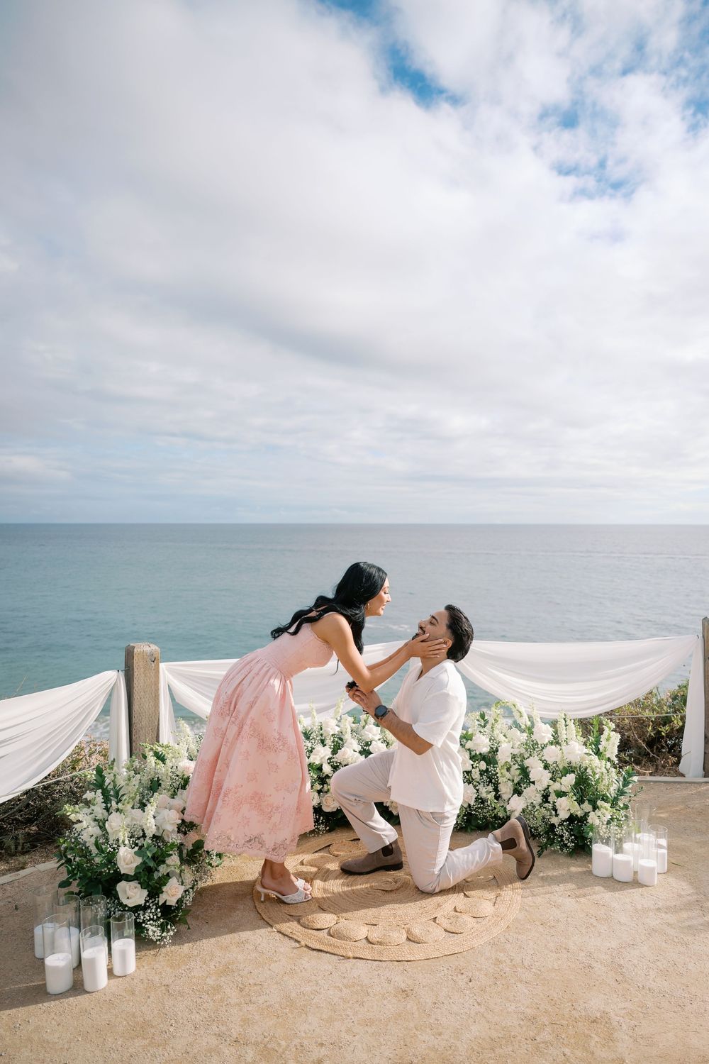Man proposes on one knee to woman in pink dress at ocean overlook decorated with white flowers and candles.