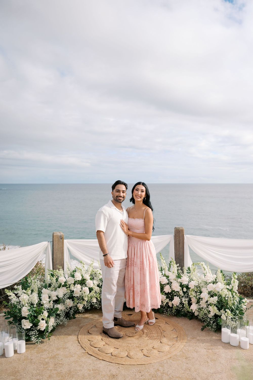 Couple posing at an oceanfront proposal setup with white floral arrangements, candles, and draped fabric backdrop.