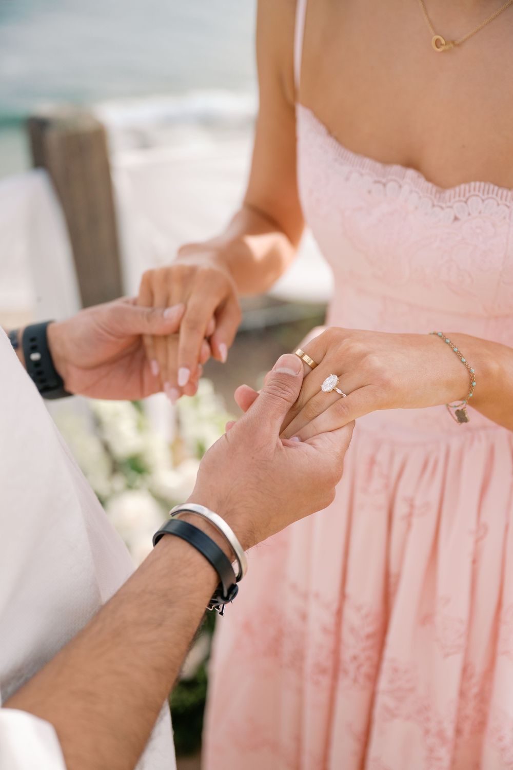 Couple exchanging rings during beach wedding ceremony, bride in pink lace dress with diamond ring.