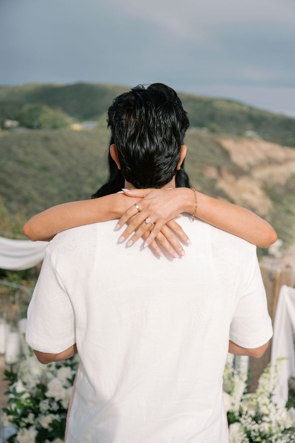 Couple embracing at outdoor wedding ceremony with scenic green hills in the background, bride's hand showing engagement ring.